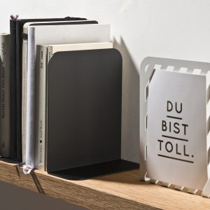 Buchstütze Solid 01 von weld & co in Schwarz, verwendet als Buchstütze für Bücher und Buchstütze Grid 01 von weld & co in Weiß, verwendet als Bilderrahmen für Fotos und Postkarten.